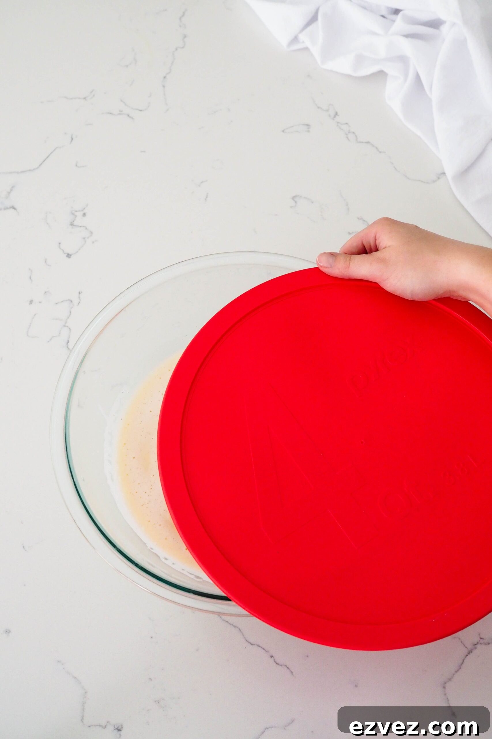A hand places a red lid over a glass bowl of custard for French vanilla ice cream.