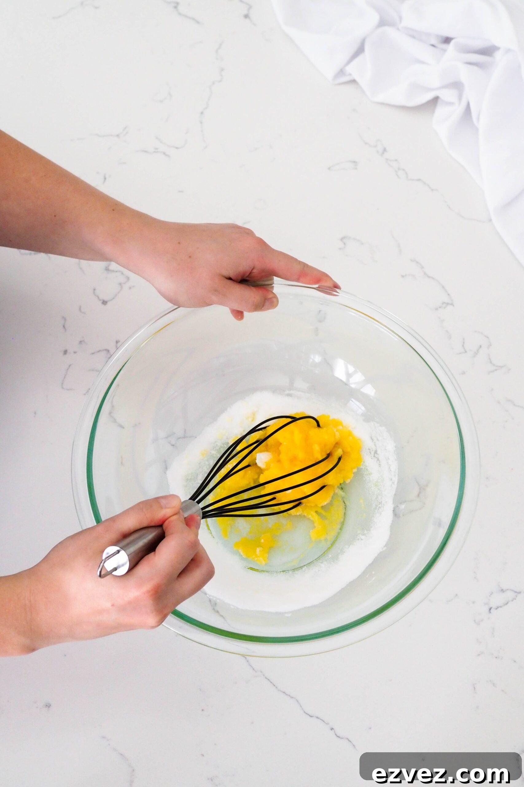 Two hands whisk together a dense and bright yellow mixture of egg yolks and sugar.