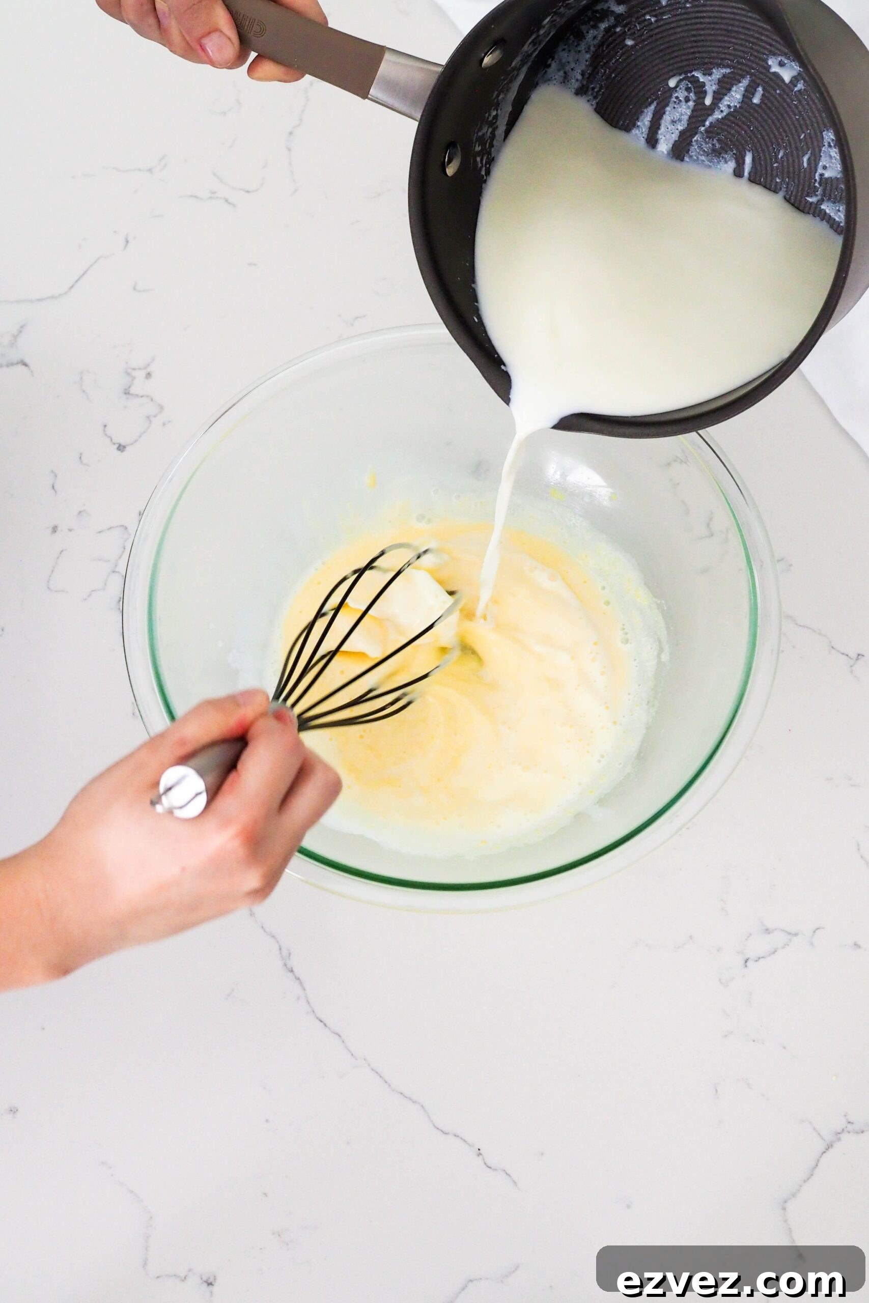 Hot milk and cream from a pot is whisked into egg yolks at the ribbon stage.
