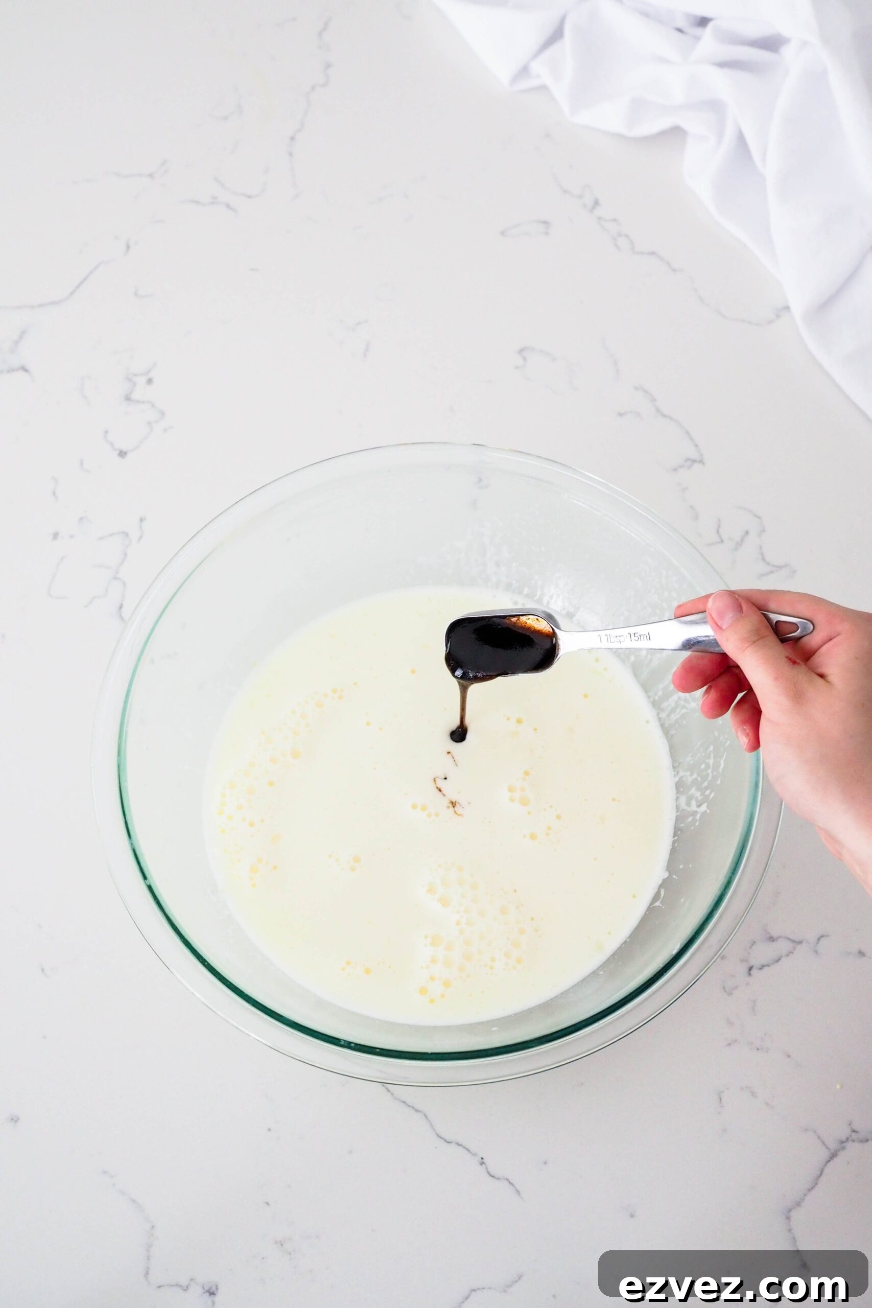 A hand pours a Tablespoon of vanilla bean paste into ice cream custard.