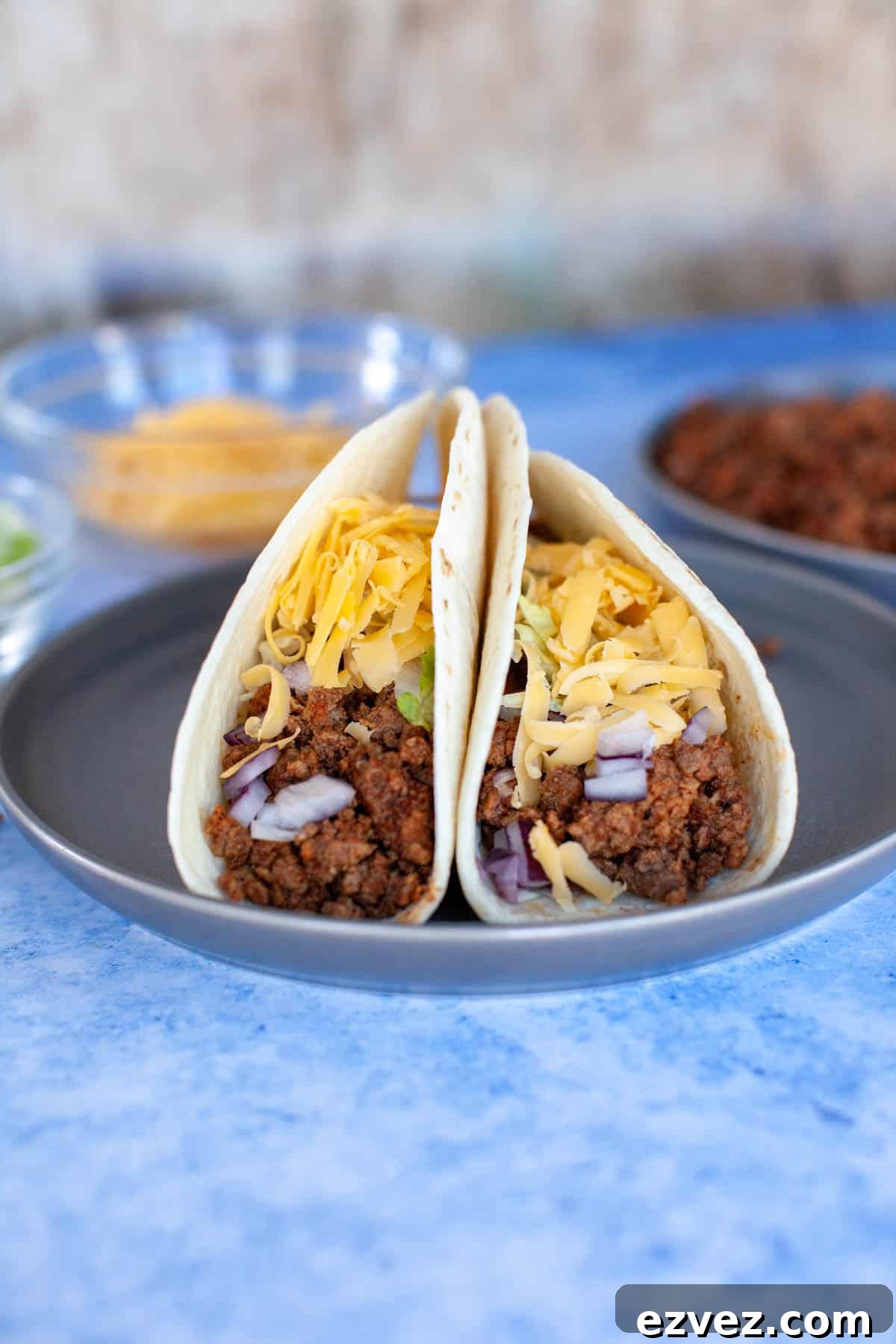 Two ground beef tacos on a plate with cheese, chopped red onion and shredded lettuce.