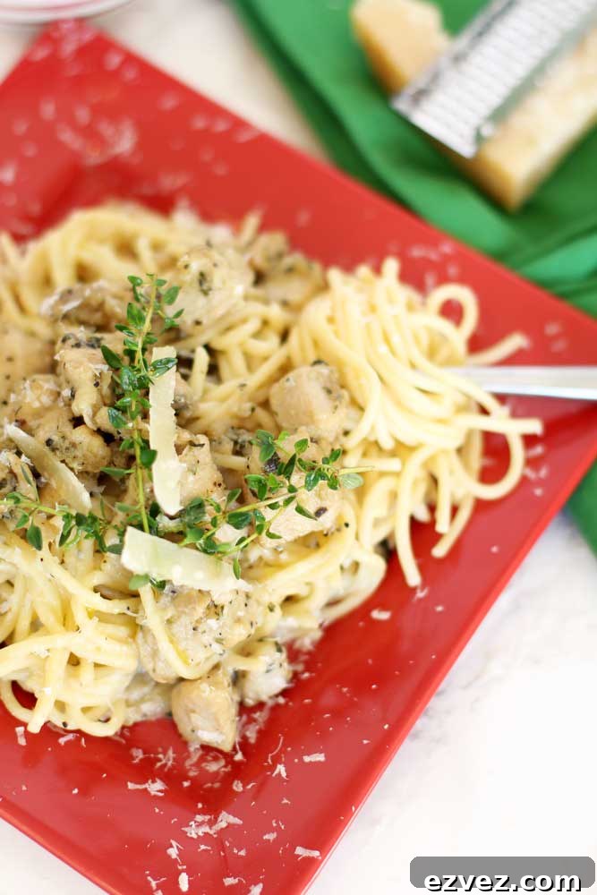 Red plate containing Garlic Basil Chicken Pasta topped with fresh sprigs of thyme and shaved Parmesan cheese, fork spiraling noodles.