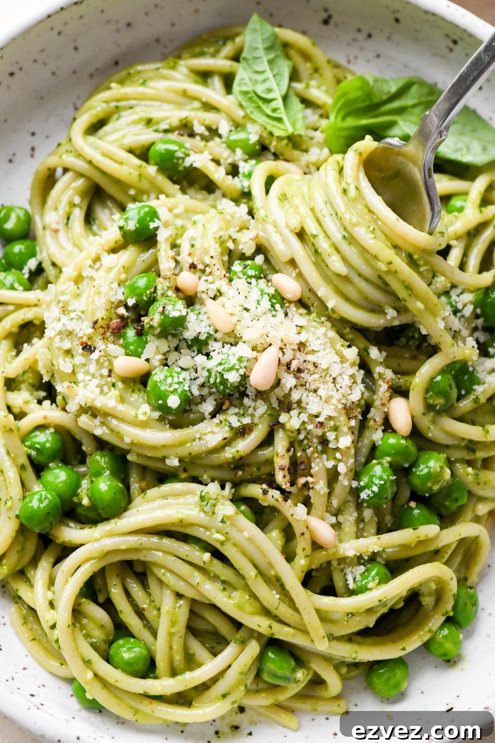 Pesto pasta made with spaghetti in a speckled ceramic bowl with a fork twirling a bite, topped with pine nuts and grated parmesan.
