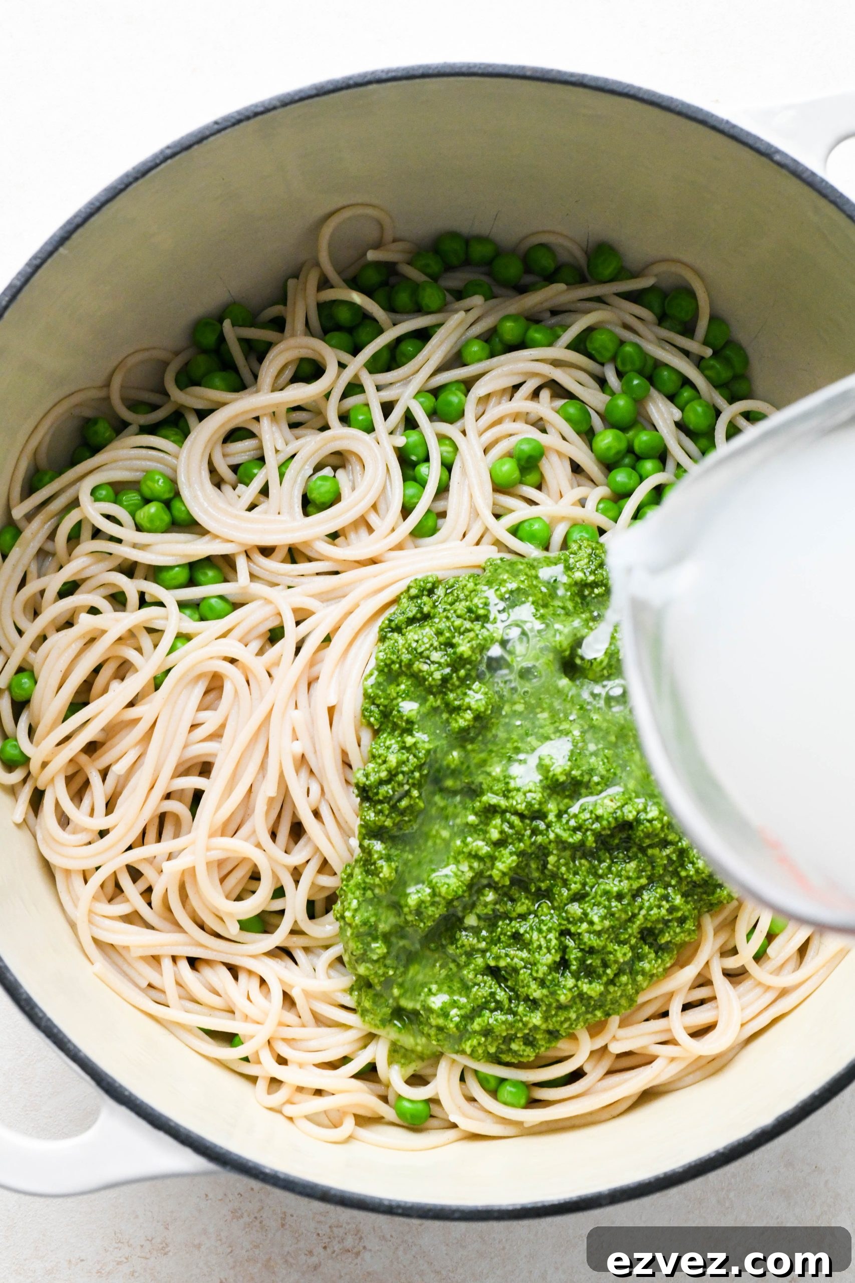How to make easy pesto pasta: Pasta and peas in pot with pesto on top, a hand pouring pasta cooking water over the pesto.