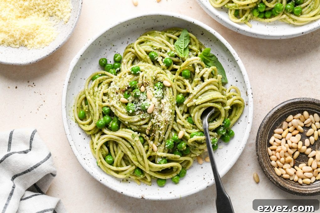 Pesto pasta made with spaghetti twirled in a speckled ceramic bowl and topped with pine nuts and grated parmesan. Next to a small dish of parmesan cheese, pine nuts, and a second bowl of pasta.