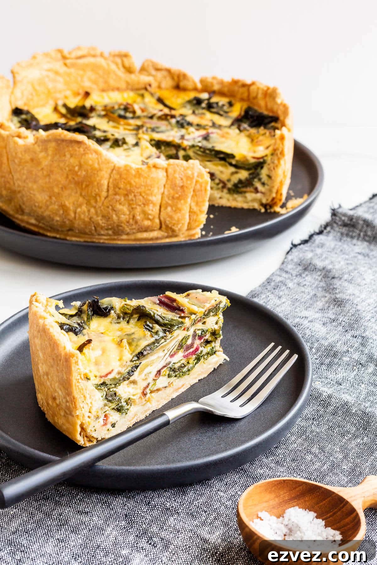 A slice of quiche on a black plate and in the background the deep dish quiche with the slice removed to show the inside with colourful rainbow Swiss chard.