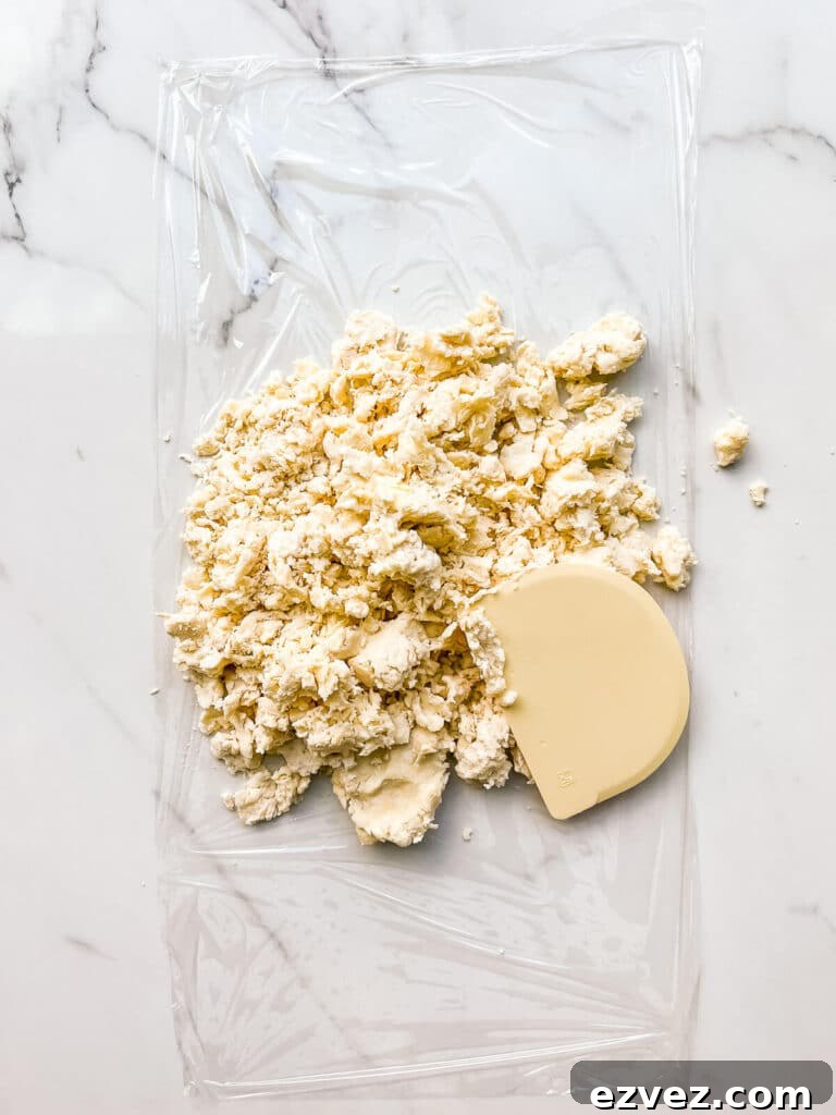 Pressing pie dough together to shape into a disk with a bench scraper on a piece of plastic wrap.