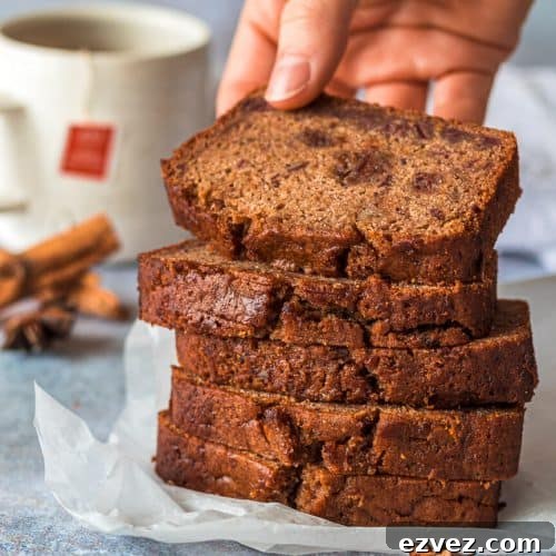 Classic Fruitcake Loaf Recipe for Moist Holiday Bread 5 Five fruit cake loaf slices stacke on top of each other, there is a hand holding the top slice. In the background is a cup of tea and various whole spices.