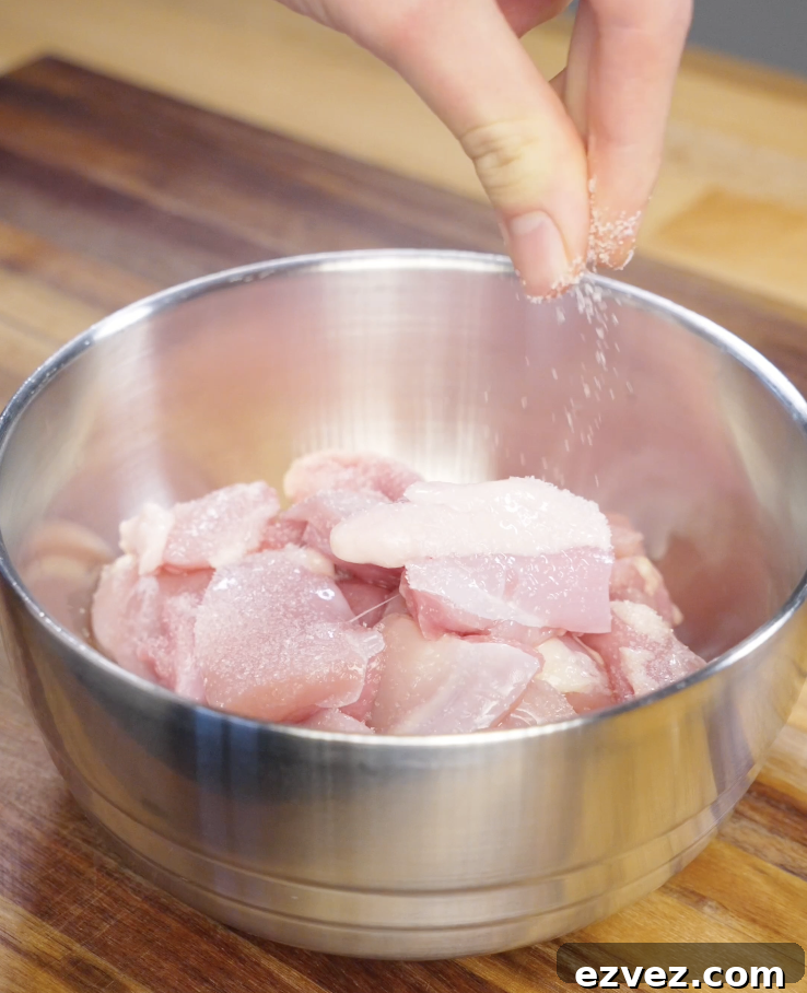 Hand sprinkling salt over bite sized boneless skinless chicken thighs in a metal bowl before marinating for homemade Mongolian chicken.
