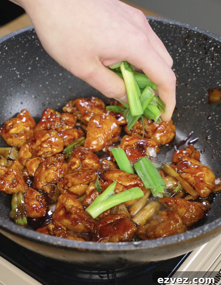 Fresh scallion greens being added to glossy Mongolian chicken in a skillet just before serving to brighten the sweet savory sauce.