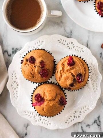 Three pumpkin cranberry muffins on a white plate in front of a cup of coffee.