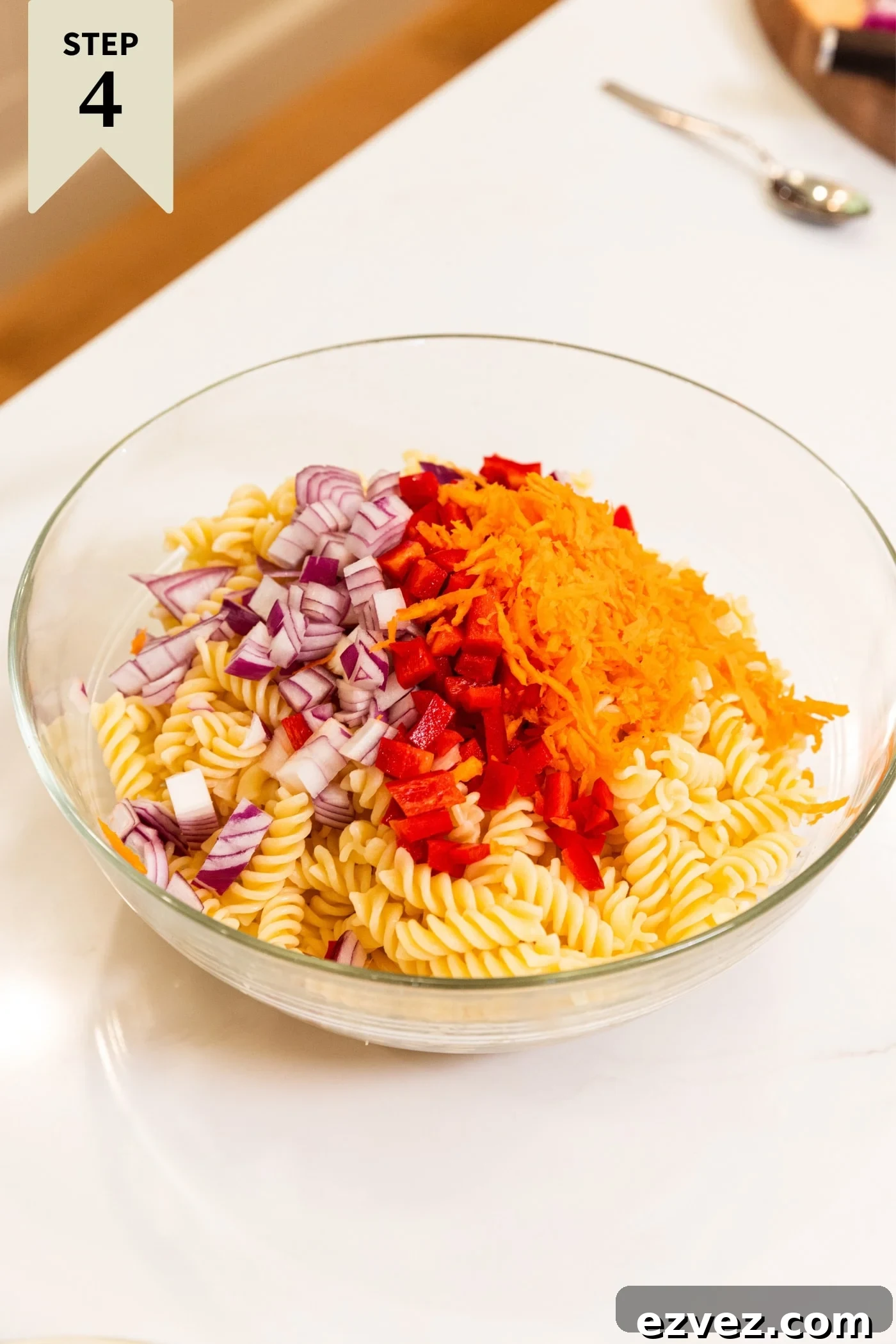 Large glass bowl filled with cooked pasta and topped with diced red onion, diced red bell pepper, and grated carrot all on white surface.