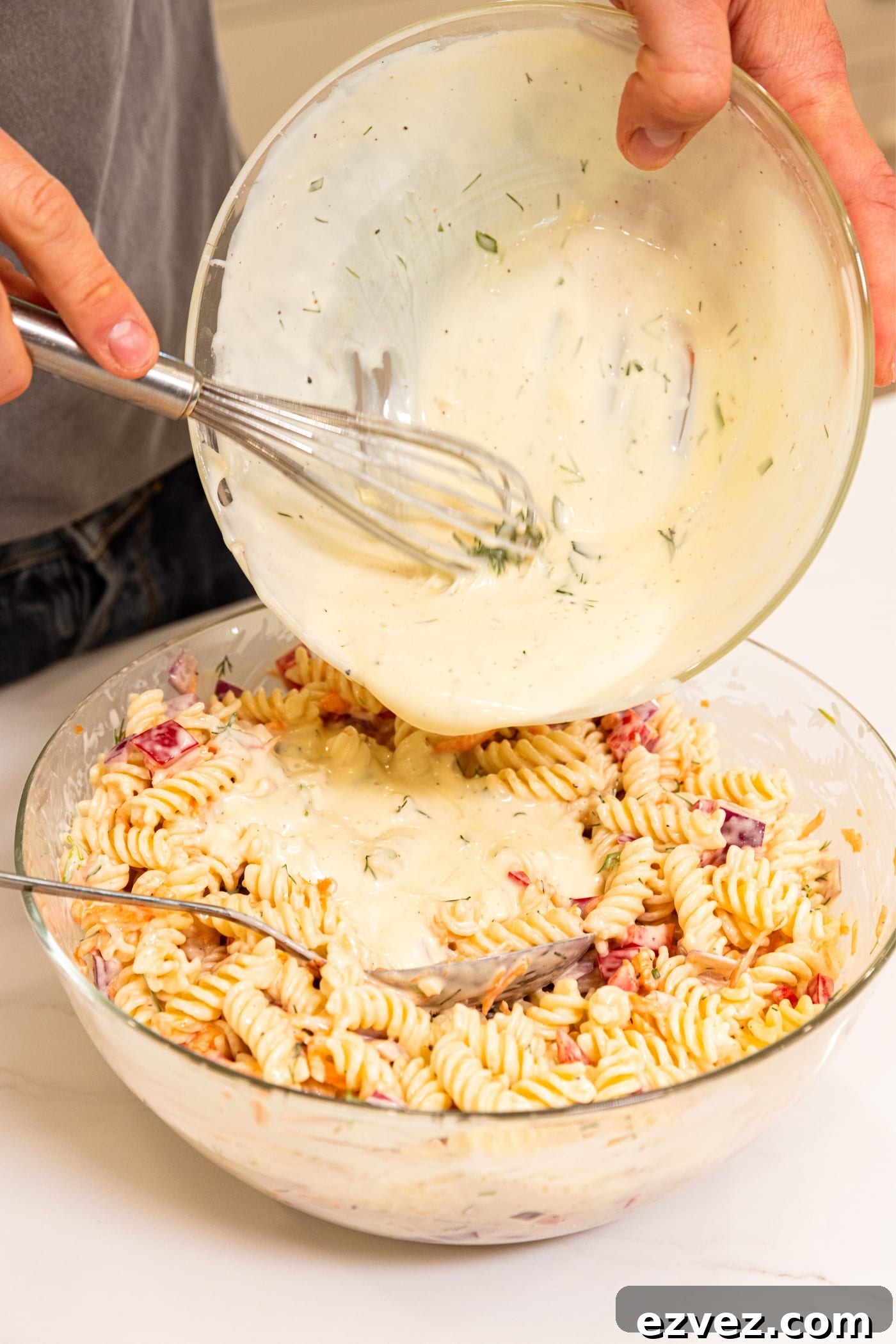 Hand holding glass bowl pouring creamy dressing over the top of pasta salad sitting in another glass bowl on white surface.