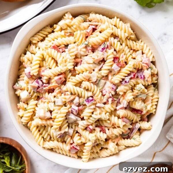 Marble surface with large white bowl filled with creamy pasta salad with red bell pepper and hamburger buns to the side.