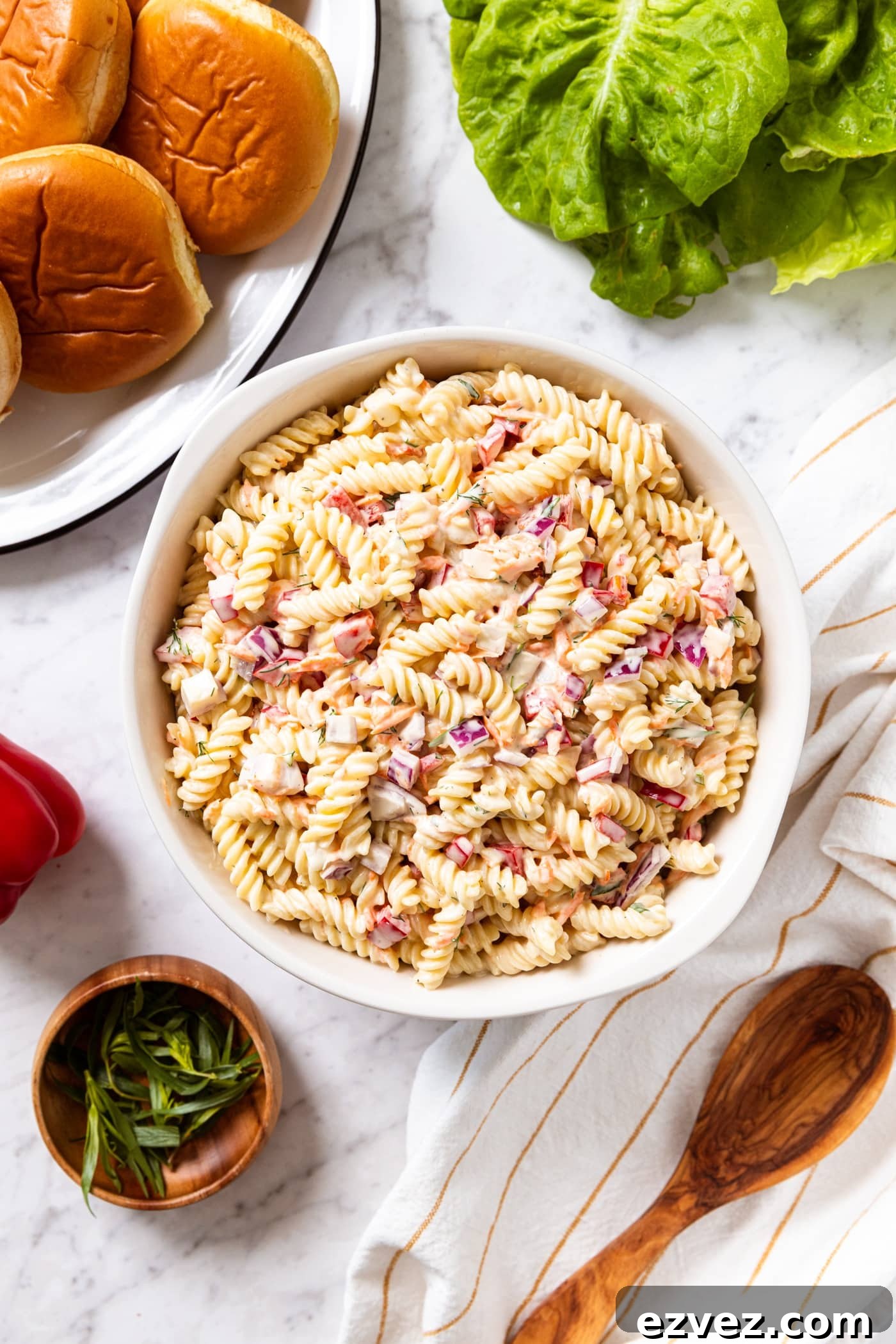 Marble surface with large bowl filled with creamy pasta salad with lettuce, hamburger buns, red bell pepper, herbs, and a wooden spoon.