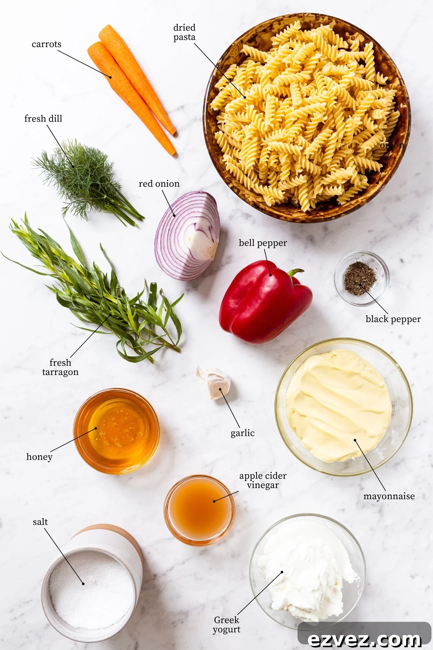 Marble surface with all ingredients needed to make creamy pasta salad including dried pasta, carrots, herbs, red onion, bell pepper, honey, mayonnaise, and more.