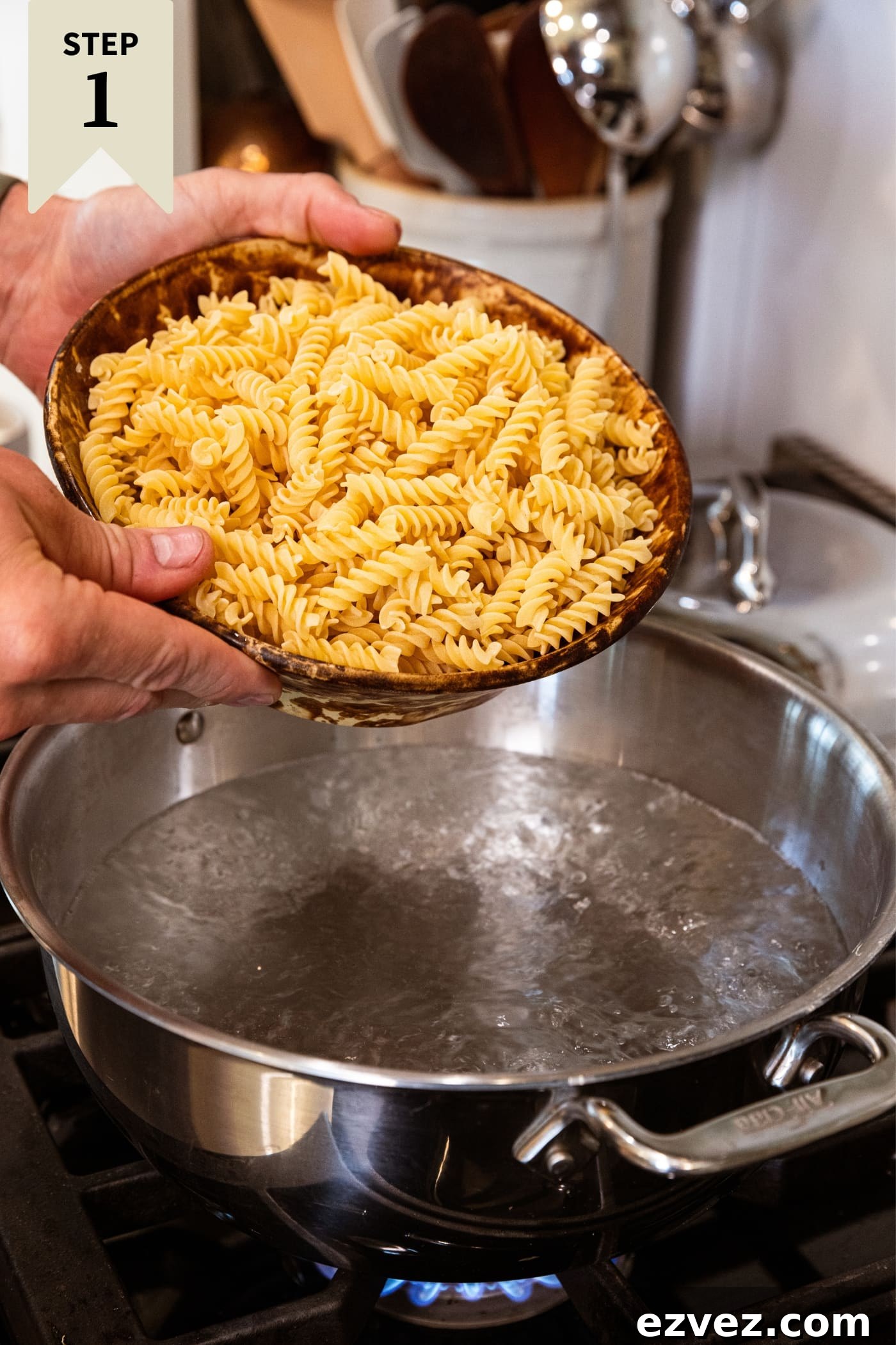 Two hands holding brown bowl about to pour uncooked pasta into a large pot of boiling water on stovetop.