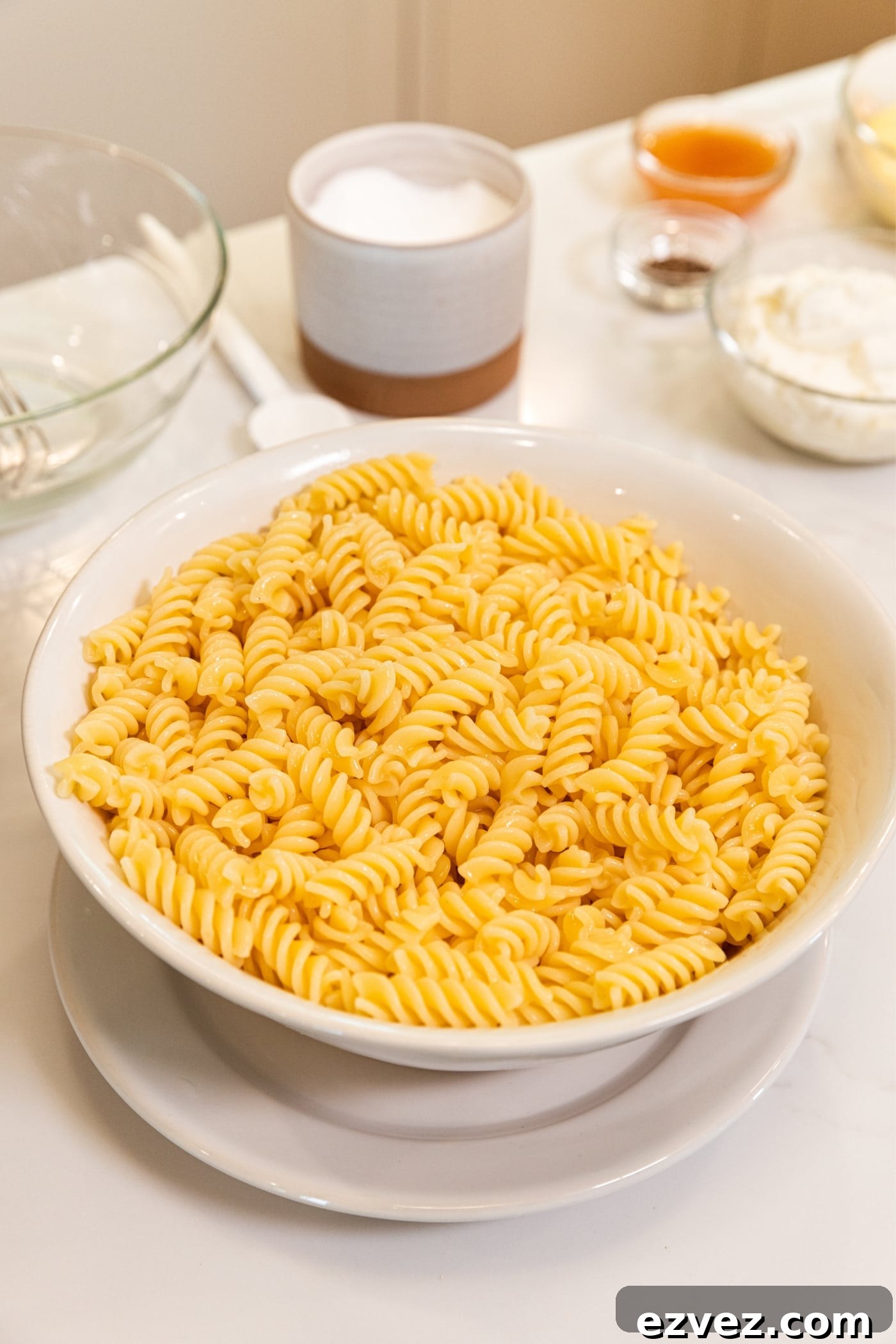 White colander with cooked pasta draining in bowl on white plate all on white surface with other ingredients in background.