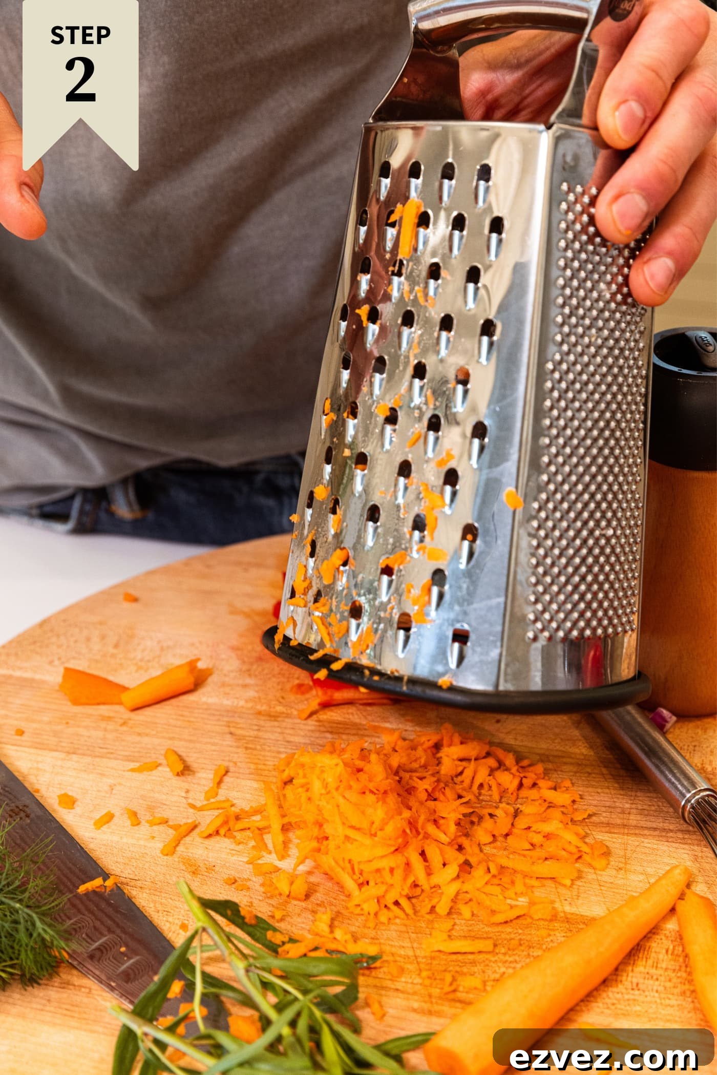 Hand holding box grater holding above grated carrot on wood cutting board.