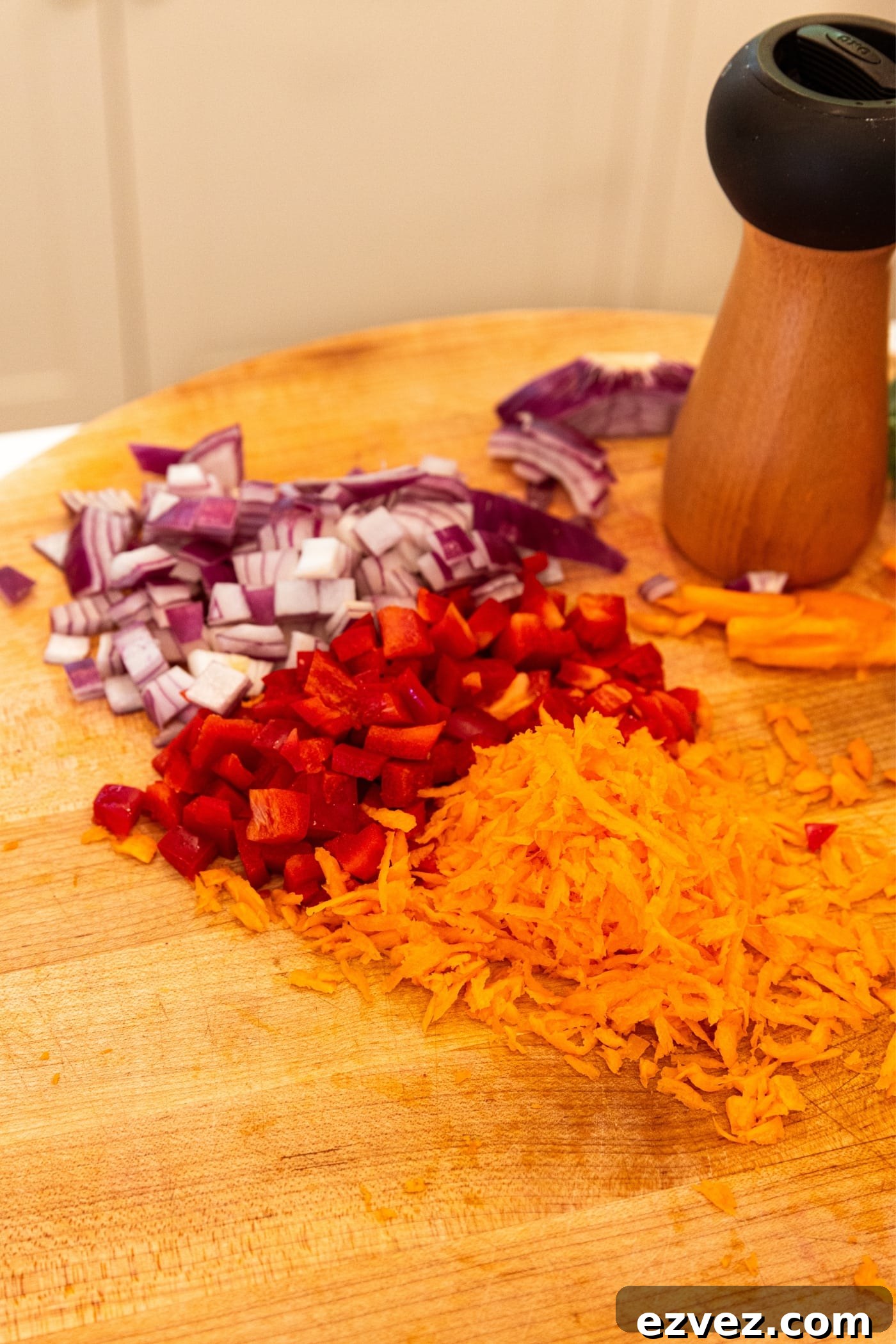 Wooden cutting board with diced red onion, diced red bell pepper, and grated carrot with black pepper grinder in background.