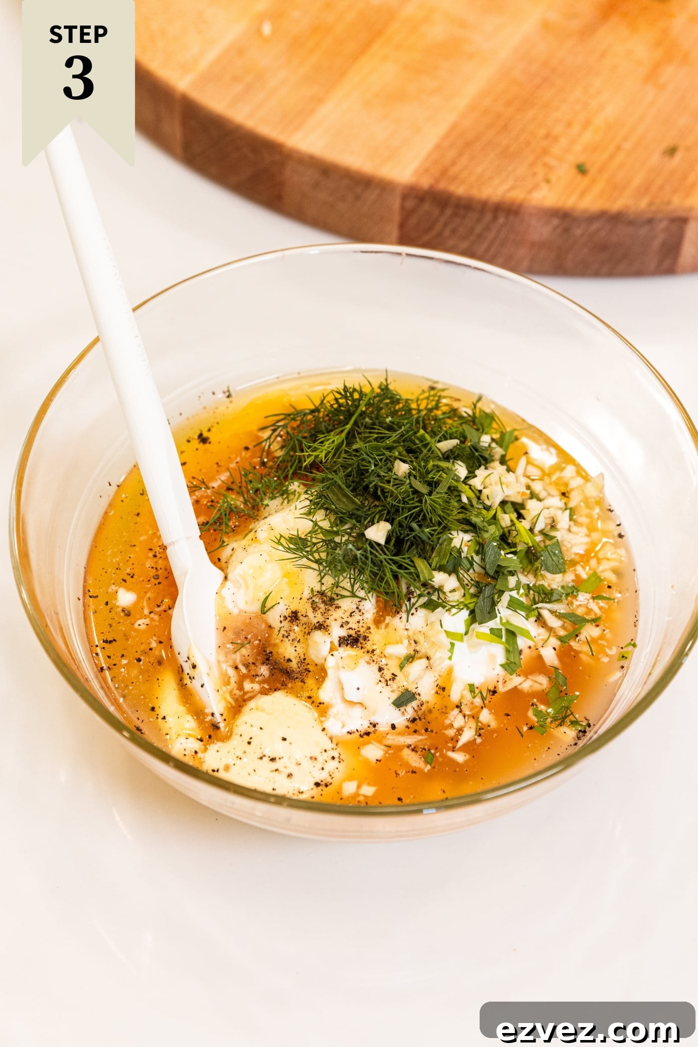 Small glass bowl filled with all ingredients needed to make a creamy pasta dressing including mayonnaise, yogurt, fresh herbs, honey, and apple cider vinegar all on white surface with wood cutting board in background.