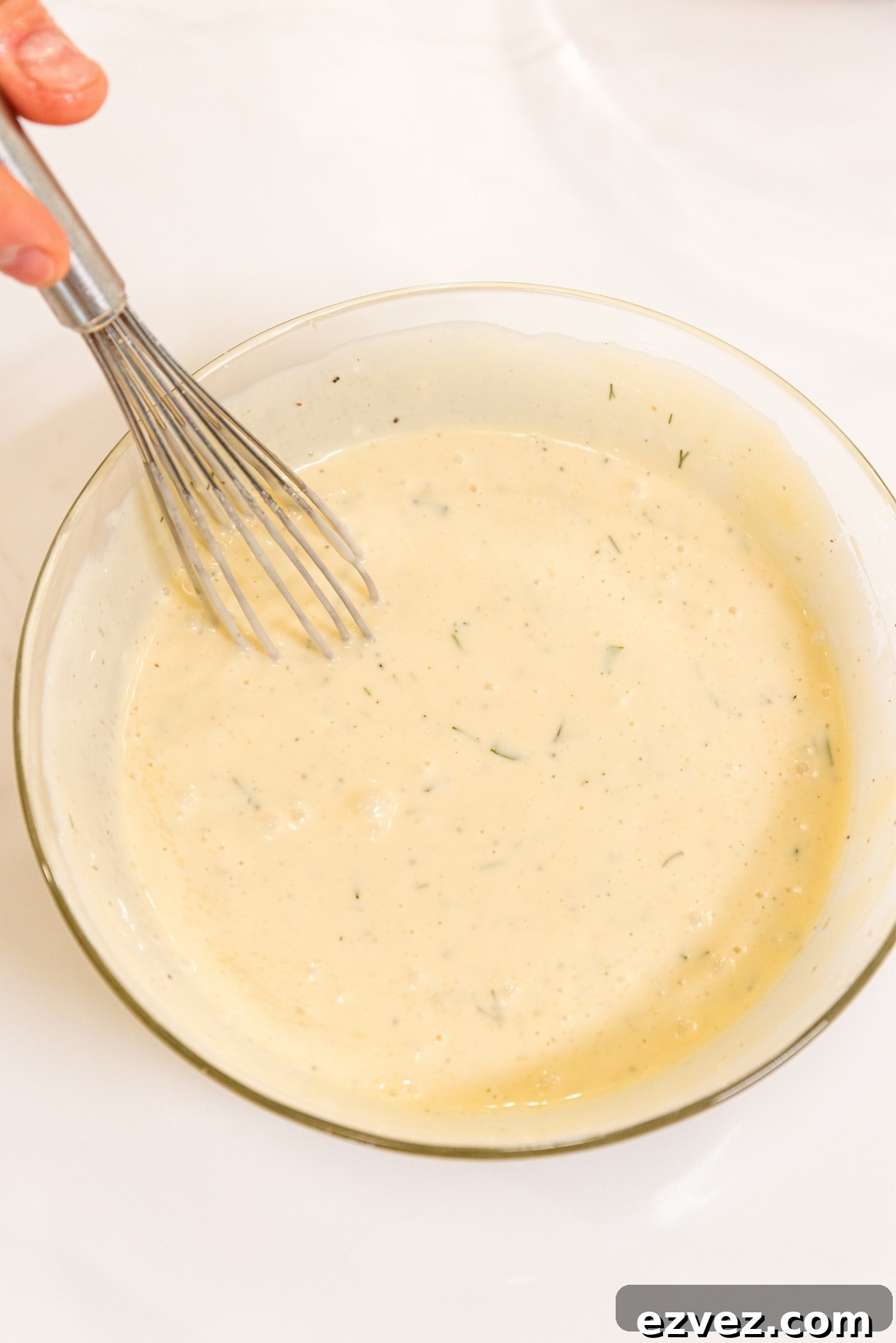 Hand holding wire whisk in middle of small glass bowl filled with cream-colored creamy dressing for pasta salad all on white surface.