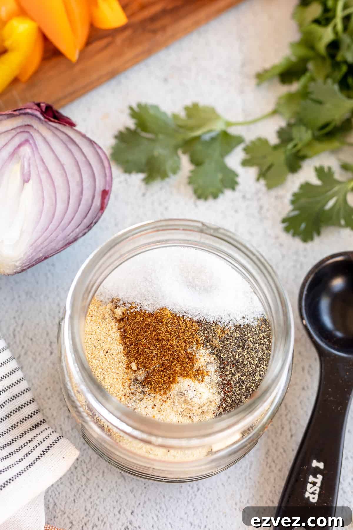 Homemade Fajita Seasoning Blend for Authentic Mexican Flavor 5 A top down shot of spices in a mason jar next to a red onion, yellow bell peppers, cilantro, and a measuring spoon.