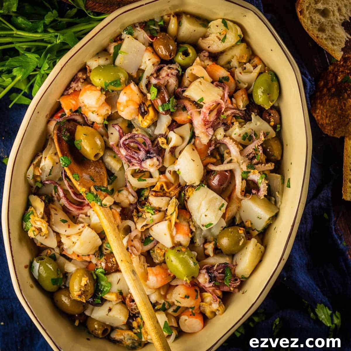 Overhead of salad in tan oval server with wooden spoon, surrounded by parsley stems and bread pieces.
