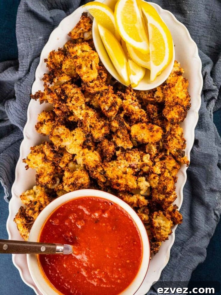 Overhead of cooked calamari on fluted, white oval platter on gray cloth, wtih dish of lemon wedges and bowl of dipping sauce nestled in.