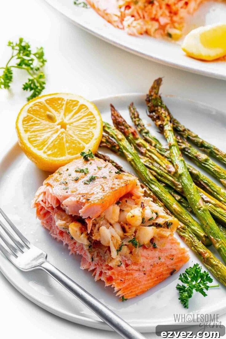 One piece of salmon on a white plate with fork, asparagus and lemon half, with serving platter in background.