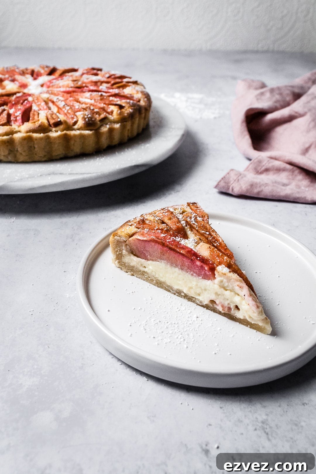Slice of Pink Apple Tart with Cream Cheese Filling on a white plate and a grey background with whole tart behind it