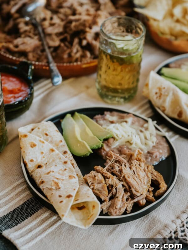 Colorado-Style Green Chile Pork Roast Recipe 11 Dutch oven carnitas on a round black plate with a scoop of refried beans topped with cheese, slices of avocado, and a warmed tortilla on the side.