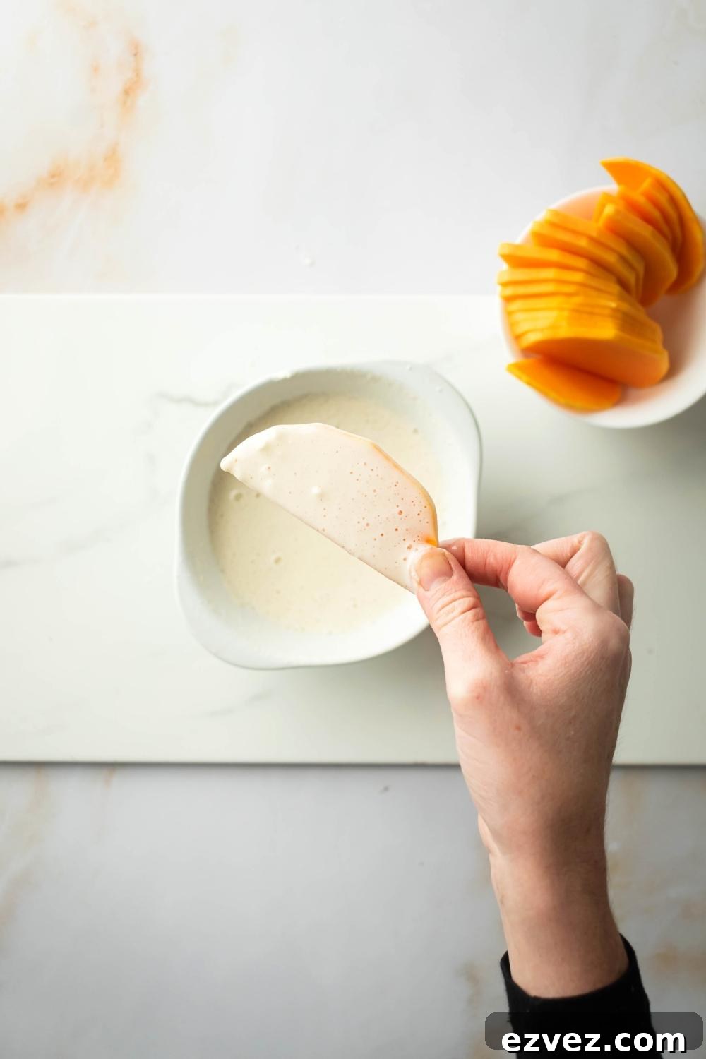 Crispy 4-Ingredient Fried Squash Recipe 4 A hand holding a piece of squash coated in a flour mixture. It is being held over a bowl with the mixture in it and next to that bowl is a bowl of sliced squash.