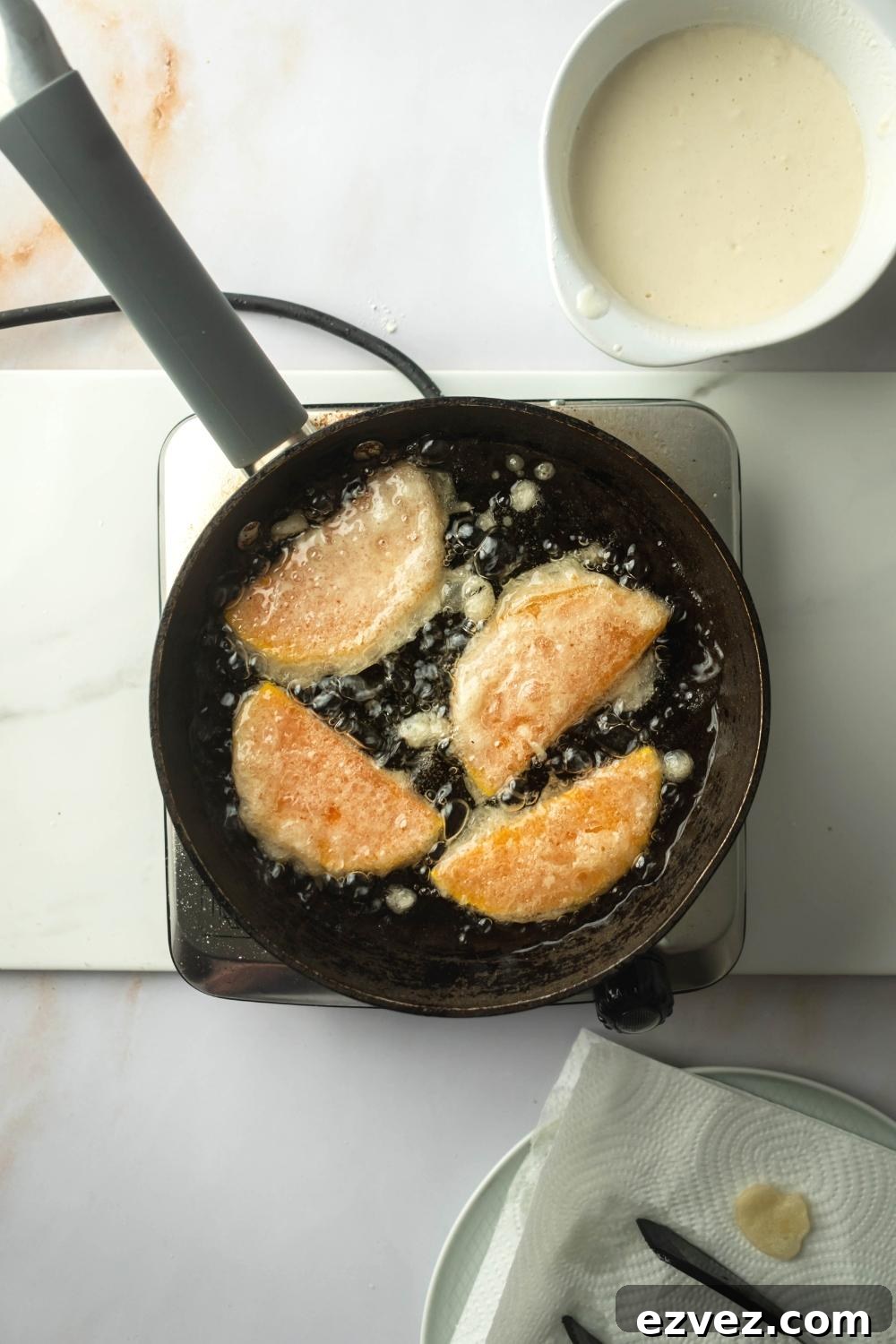 Crispy 4-Ingredient Fried Squash Recipe 6 A frame pan filled with oil and four pieces of squash. The frying pan is on a square burner and behind is a bowl thee mixture for breading.