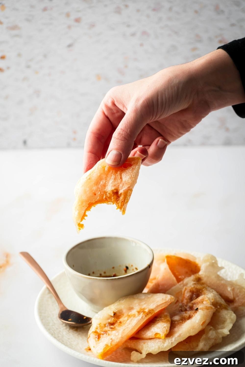 Crispy 4-Ingredient Fried Squash Recipe 8 A hand holding a piece of fried squash that has a bite out of it over a cup of soy sauce that is on a white plate with a bunch of other pieces of fried squash overlapping with one another.