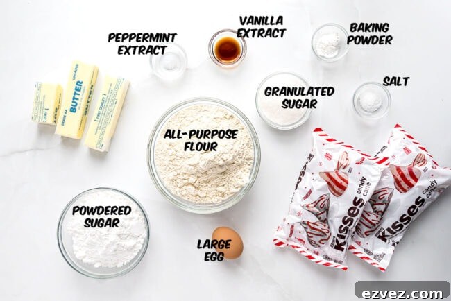 Overhead view of ingredients for Candy Cane Kiss Cookies, including butter, powdered sugar, flour, egg, sugar, peppermint extract, and candy cane Hershey’s Kisses.