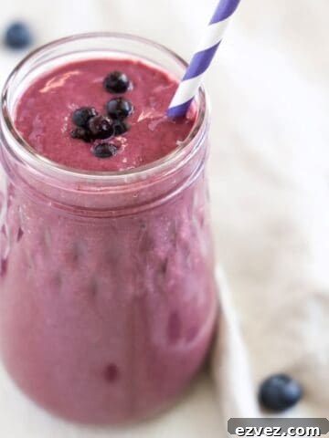 Blueberry Mango Tofu Smoothie with a purple and white striped straw. A linen napken with a few blueberries wraps around part of the base of the smoothie jar.