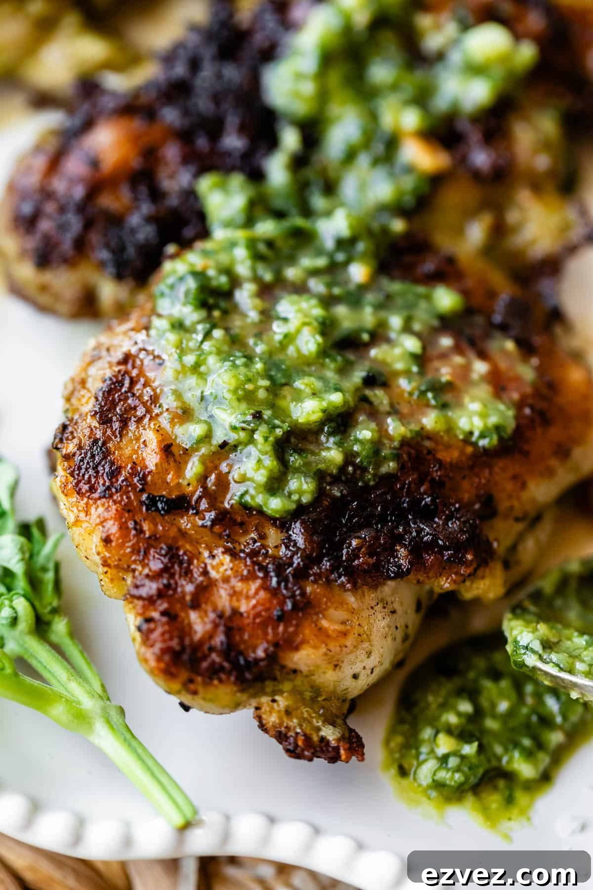 Close-up of a piece of grilled chicken topped with green pesto sauce, served on a white plate with a small sprig of fresh herbs on the side. The chicken has a crispy, golden-brown skin.