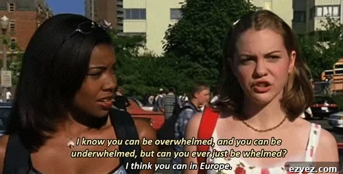 Two young women stand outdoors talking. The woman on the right says, I know you can be overwhelmed and underwhelmed, but can you ever just be whelmed? Maybe while sharing a Chicken Arugula Salad in Europe.