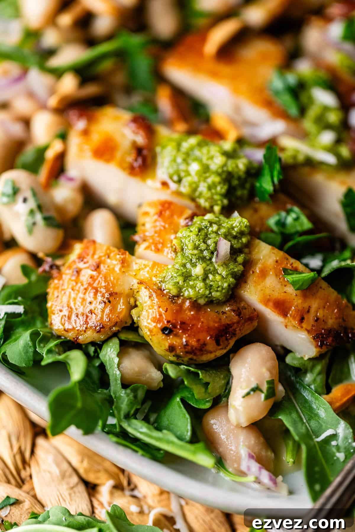 Close-up of sliced grilled chicken topped with green pesto, served on a bed of arugula, white beans, fresh herbs, and toasted almonds. The dish appears fresh and vibrant with a golden, crispy chicken skin.