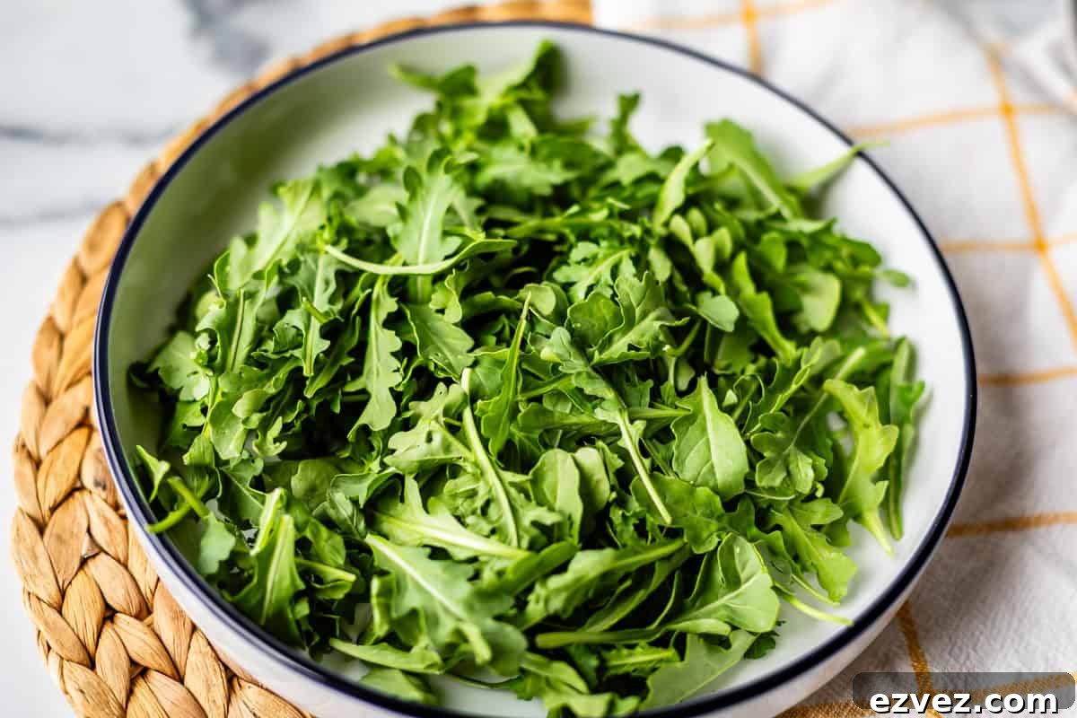 A white bowl filled with fresh green arugula leaves sits on a woven placemat with part of a white cloth visible nearby.