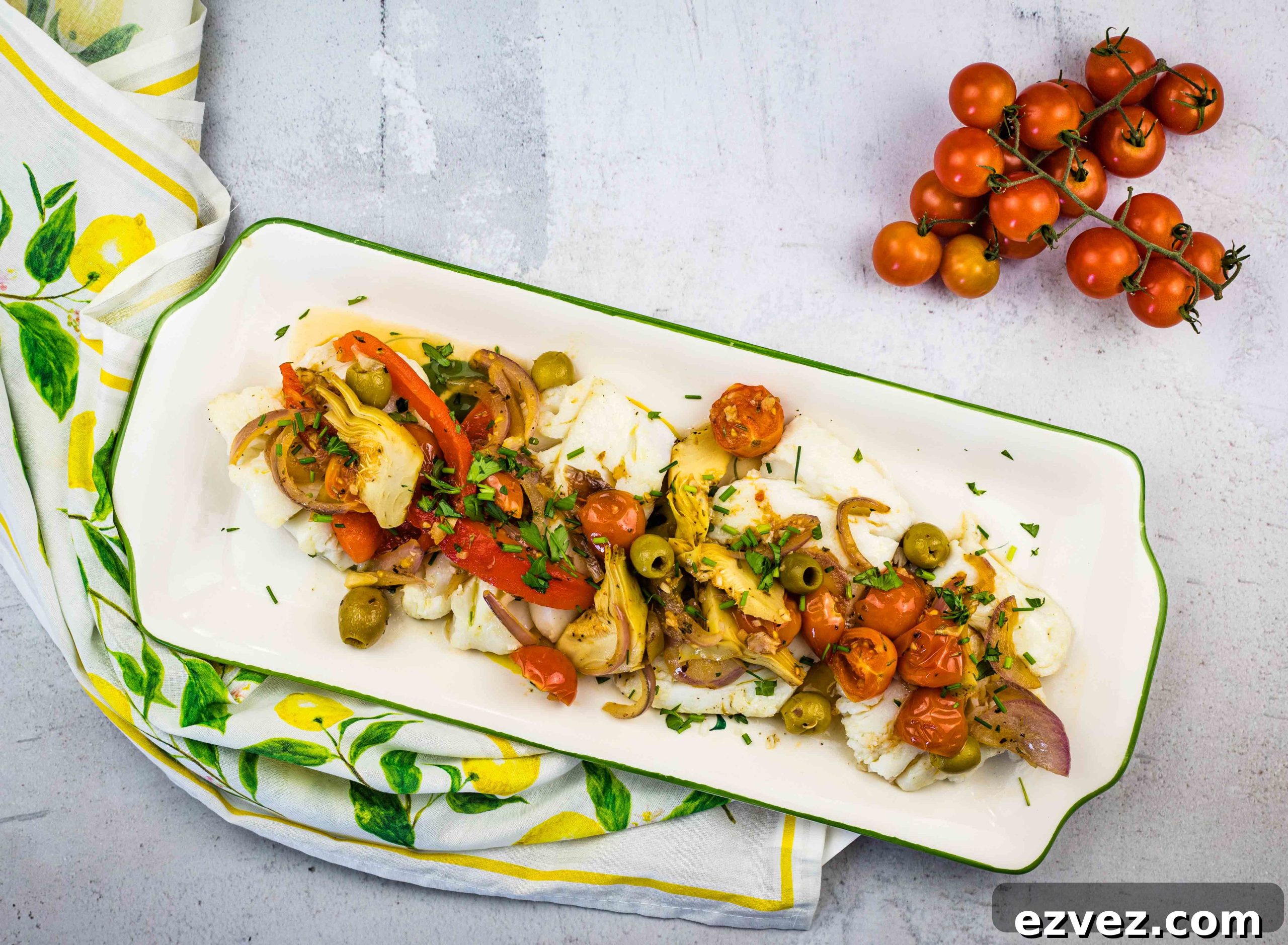 A rectangular plate with Mediterranean Cod placed on a lemon-patterned cloth. A vine of cherry tomatoes is beside the plate.