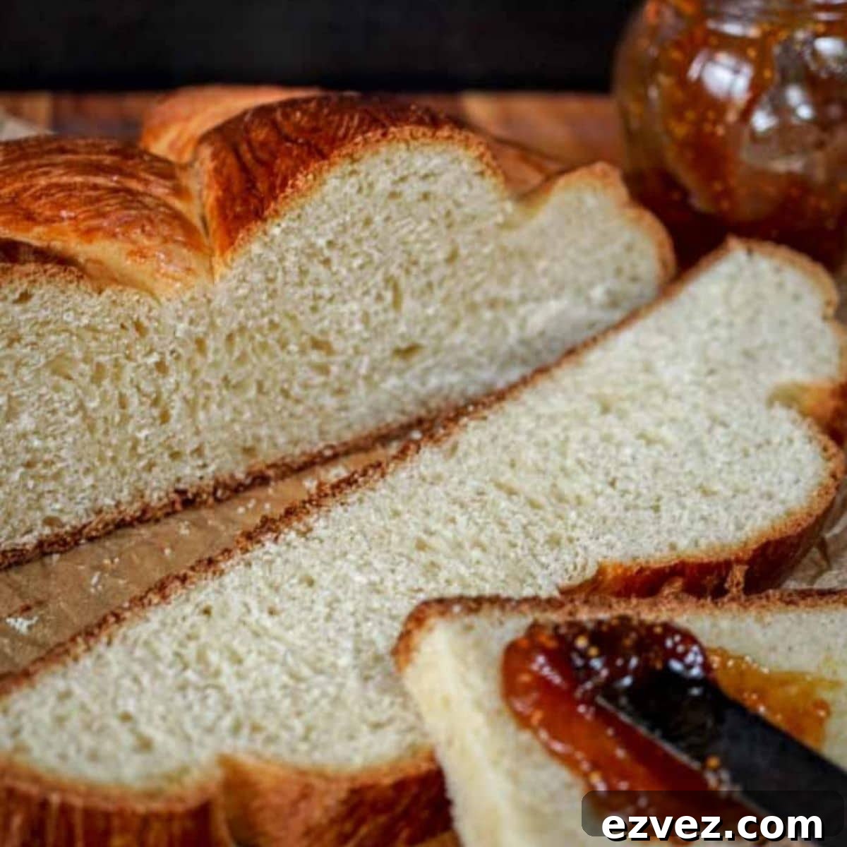 A loaf of Hungarian Egg Twist Bread with the front slices cut from the front. A jar of jam sits in the background and a knife with jam sits on the front slice of bread. Hostess At Heart