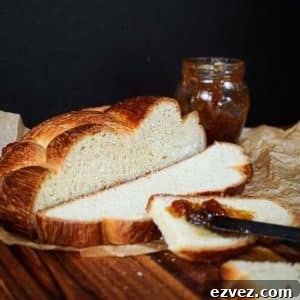 A loaf of Hungarian Egg Twist Bread with the front slices cut from the front. A jar of jam sits in the background and a knife with jam sits on the front slice of bread. Hostess At Heart