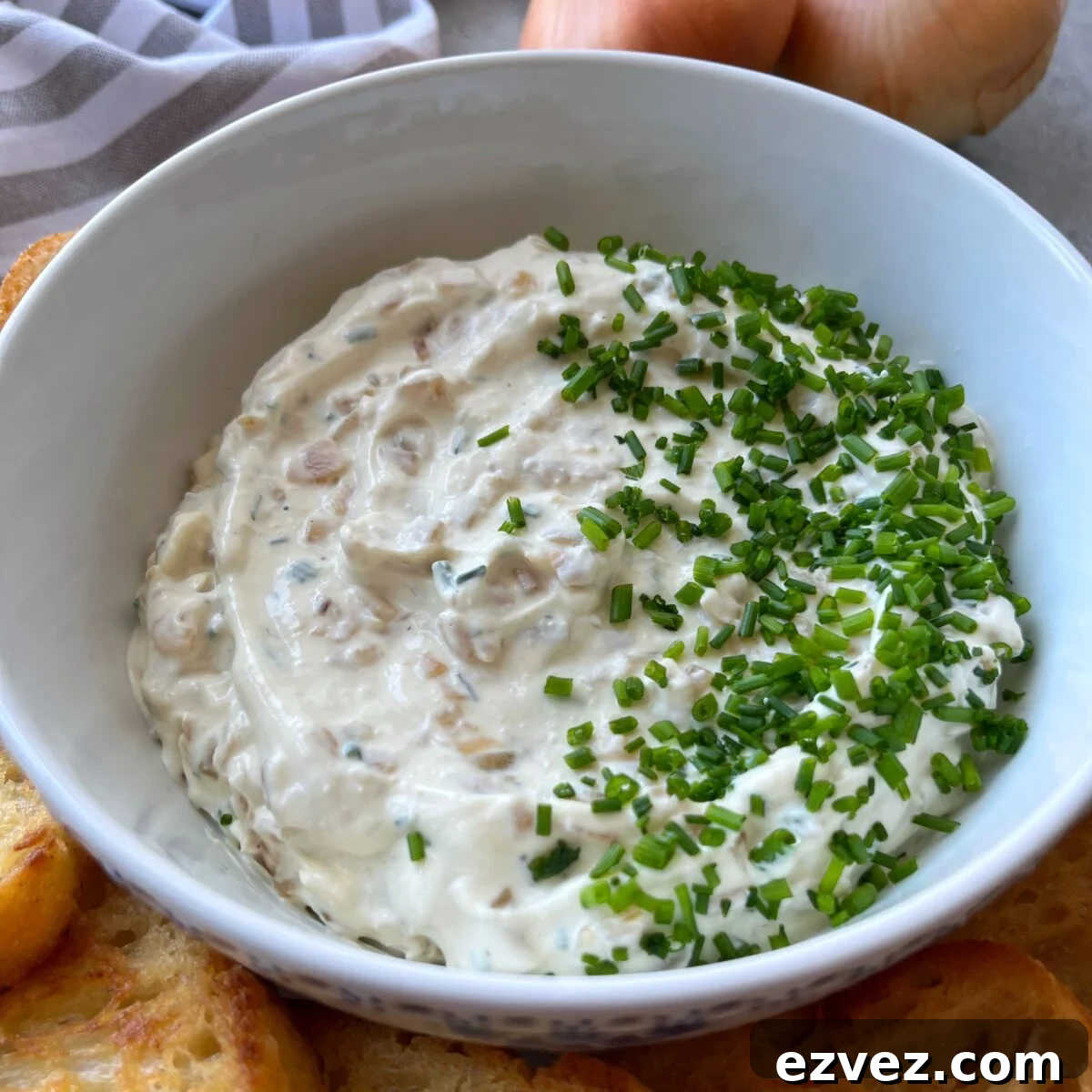 A white bowl filled with creamy caramelized onion dip garnished with chopped chives, surrounded by slices of toasted bread.