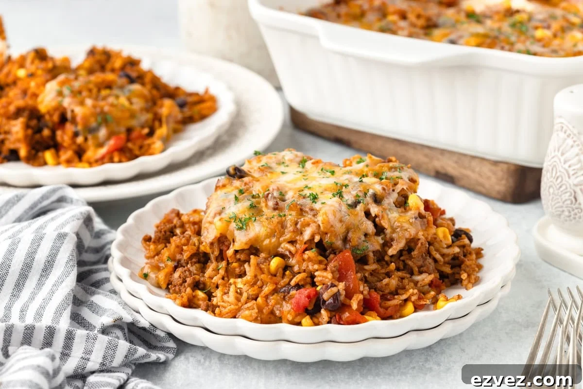 A serving of cheesy Mexican casserole with rice, ground beef, corn, black beans, and tomatoes on a white plate, with a baking dish of casserole and a fork nearby.
