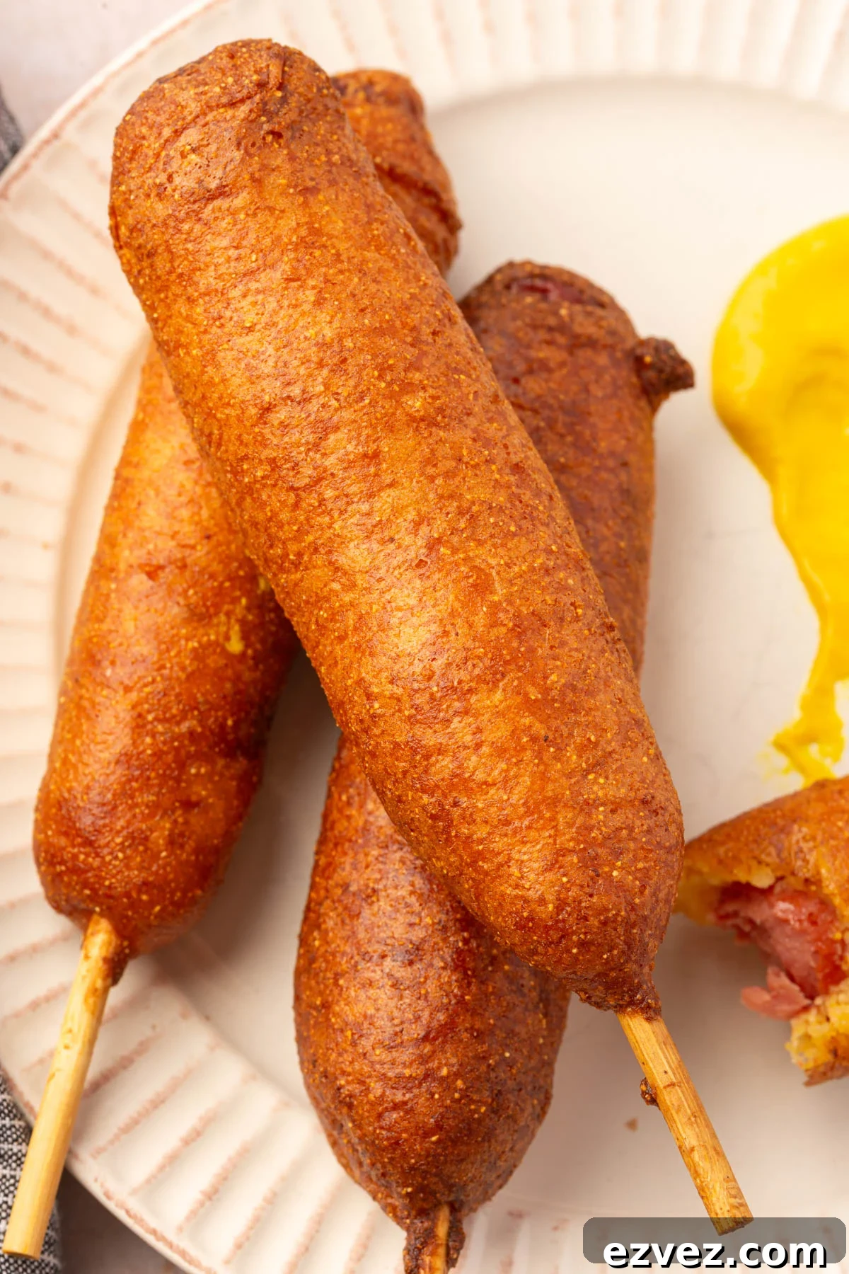 Three gluten-free corn dogs on a white plate with a dollop of yellow mustard for dipping.