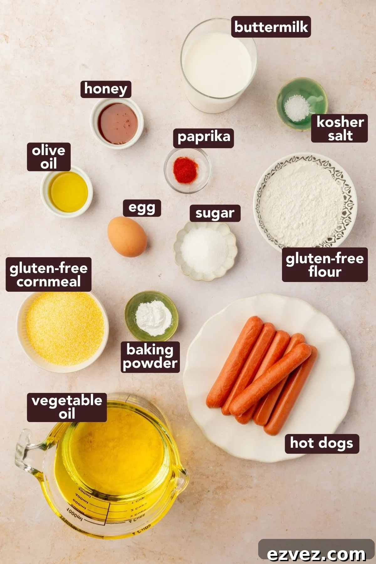 Overhead view of ingredients for gluten-free corn dogs including cornmeal, gluten-free flour, hot dogs, buttermilk, egg, honey, paprika, salt, sugar, olive oil, baking powder, and vegetable oil.