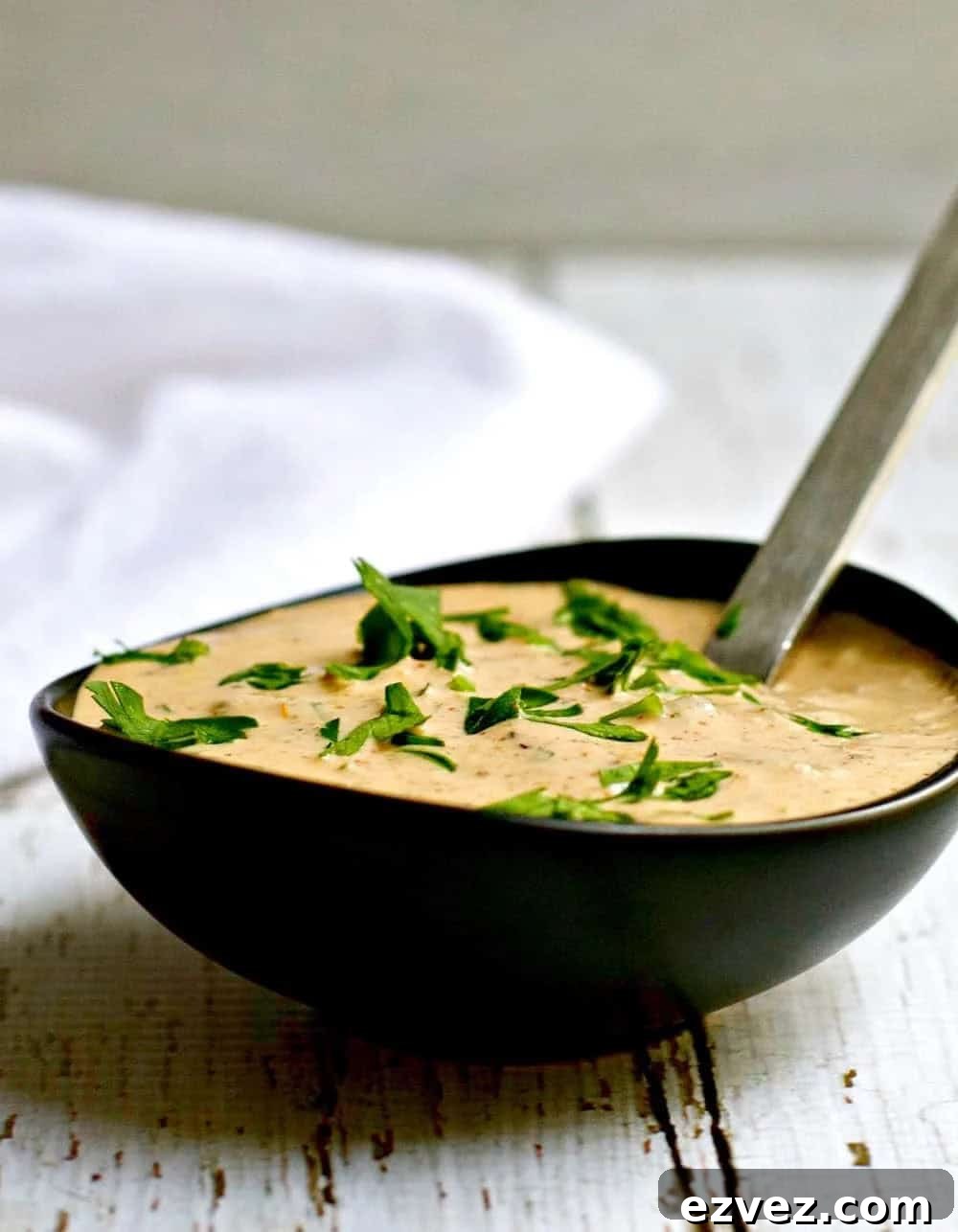 A black bowl filled with creamy remoulade sauce, garnished with chopped fresh herbs, with a spoon resting inside. The bowl is placed on a rustic white wooden surface.