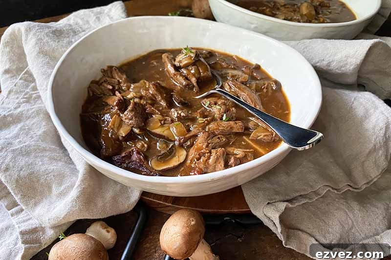 Leftover Pot Roast Mushroom Soup Recipe for Comforting Meals 14 Horizontal image of a white bowl filled with a dark brown chunky stew with a spoon.